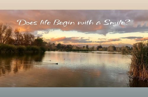 A peaceful lake reflecting the golden hues of sunset, with gentle clouds and trees in the background. Ducks swim across the calm water. The text on the image reads: “Does life begin with a Smile?”