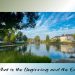 A calm lake reflecting houses, trees, and a blue sky with soft white clouds, framed by greenery and a quiet atmosphere of stillness.